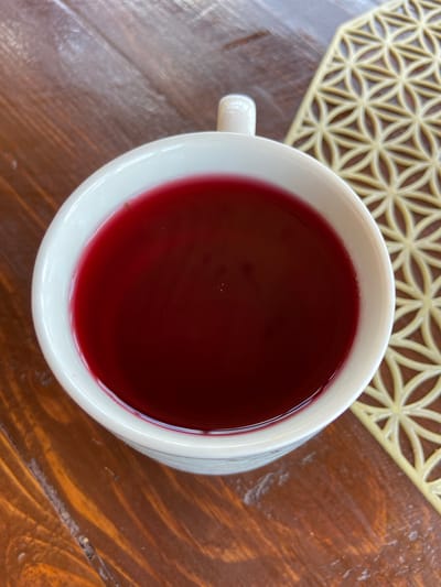 A close-up, top-down shot shows a white mug filled with a deep red liquid, likely tea or juice, on a wooden table. To the right of the mug is a light yellow, geometrically patterned placemat. The scene is set in the city of Krasnomakskoye rural settlement, Ukraine.