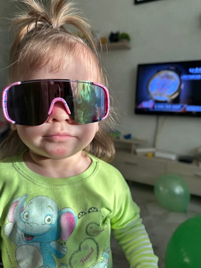 A young child is captured in a close-up, sideways portrait, set indoors in Uva, Russia. The child, likely a toddler or preschooler, has light blonde hair pulled back in a ponytail and is wearing oversized, reflective sunglasses with a pink frame accented with blue and pink specks. The lenses of the sunglasses show a distorted reflection of the surroundings, possibly including a window.
The child is wearing a light green long-sleeved t-shirt featuring a cartoon elephant design in blue, pink, and grey, along with other decorative elements. The word "Friend" is visibly embroidered or printed within a heart shape on the shirt's upper right side. A slight redness is noticeable on the child's upper lip area, extending towards the chin.
In the background, a television screen is visible, displaying what appears to be a graphic or map with prominent blue and red colors. Some indistinct white text or numbers are also on the screen. To the right of the child, partially visible, are two green balloons, suggesting a celebratory atmosphere. The lighting in the room appears bright, possibly from natural light, casting reflections.
