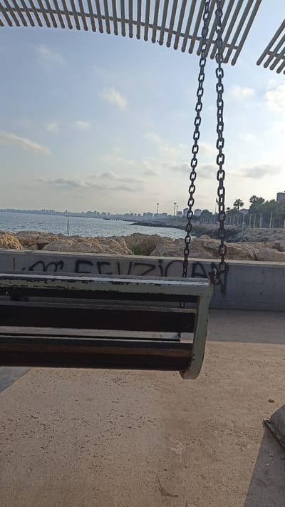 This media file captures a peaceful outdoor scene with a swing in the foreground, set against a backdrop of the sea and a distant city skyline. The swing, made of wood and suspended by metal chains, is the primary object. It hangs from a wooden structure with parallel slats, suggesting it's part of a park or recreational area.
The scene depicts a tranquil seascape with calm water stretching towards a horizon dotted with buildings. Large rocks form a barrier between the water and the paved area where the swing is located. Palm trees and more architectural structures are visible along the coastline, hinting at an urban or resort setting. The sky is a soft blue with scattered clouds, indicating fair weather. The lighting suggests it is either early morning or late afternoon.
Visible graffiti on a concrete barrier beneath the swing adds a touch of urban art to the natural and man-made elements. The graffiti appears to be in black lettering, but it's difficult to decipher fully. There's also a faint marking that looks like "ER" on the wooden slats of the swing itself.
The overall mood is one of serenity and quiet contemplation. The empty swing suggests a moment of pause, inviting relaxation and enjoyment of the view. The location, Yenişehir, Turkey, provides context for the architectural style and the presence of palm trees, consistent with a coastal Mediterranean environment. There are no people visible in the frame, emphasizing the stillness of the scene.