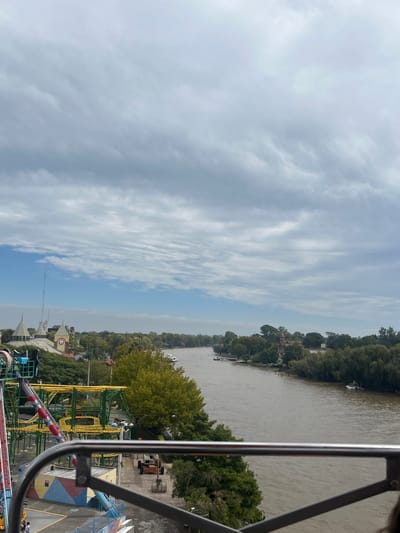 An elevated view captures a wide, brown river flowing through the city of Tigre, Argentina, under a partly cloudy sky with patches of blue. In the foreground on the left, an amusement park structure features yellow and red steel roller coaster tracks. A gray metal railing from the vantage point frames the bottom of the image. Below the ride, a paved area contains utility vehicles and colorful building facades. The left bank of the river is lined with dense green trees and shows further amusement park buildings, including structures with dome roofs and a building with a visible clock face, indicative of Parque de la Costa. Several boats are moored along this bank. The river curves slightly to the right in the mid-ground. The right bank is heavily forested, with a distinctive red-brick building featuring multiple turrets and white accents visible through the trees. More boats are present on the river, both moored and freely floating. The distant horizon appears hazy.