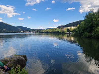 The image depicts a serene lakeside scene in Pancharevo, Bulgaria. The foreground is dominated by a calm body of water, with gentle ripples reflecting the bright blue sky and scattered white clouds. On the left bank, a person is seated on a large rock, holding a fishing rod, suggesting an activity of leisure. Beyond the water, a lush green landscape unfolds, with trees and vegetation lining the shore. In the distance, buildings and structures are visible, nestled against the backdrop of rolling hills and mountains, indicating a semi-urban or suburban setting. The overall impression is one of tranquility and natural beauty, with a hint of human presence engaged in a peaceful pastime.