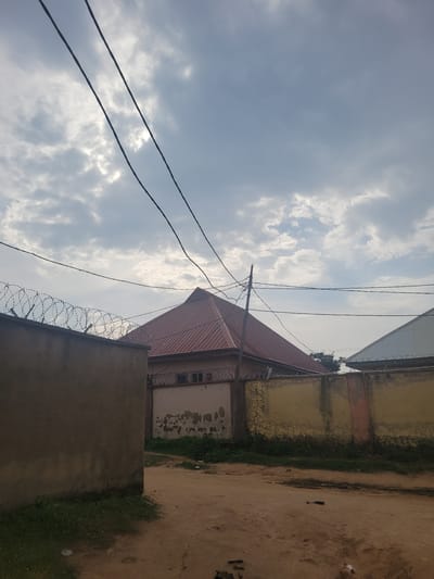 In the city of Suleja, Nigeria, an overcast sky with scattered clouds forms the backdrop for a view of residential buildings and surrounding walls. Two power lines traverse the upper portion of the frame. In the foreground, a dirt path leads past a rough, light-brown wall topped with razor wire on the left. To the right, a yellow-painted wall with patches of peeling paint and some orange staining runs along a property line. Behind the walls, a building with a red, pitched roof and a facade featuring several small, barred windows is visible. Another building with a similarly pitched, gray roof is partially obscured by the yellow wall. The environment appears to be a somewhat unkempt urban or semi-urban area, with sparse vegetation growing along the base of the walls and on the side of the path. The overall impression is one of a typical residential street in Suleja.