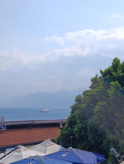 The image captures a scenic view from a slightly elevated vantage point overlooking the Mediterranean Sea, likely in Antalya, Turkey, given the architectural style of the rooftop. In the distance, hazy blue mountains form a majestic backdrop against a sky filled with soft, scattered clouds. A large, white yacht is visible on the calm blue water. In the foreground, a tiled rooftop with terracotta tiles stretches across the lower portion of the frame, suggesting a building or structure. Several outdoor umbrellas, one white and two blue, are partially visible, indicating a seating or dining area, possibly a cafe or restaurant. Lush green foliage from a large tree dominates the right side of the image, with its branches and leaves creating a dense canopy. A portion of a sign with the text "NESİ" is visible on the building's facade. The overall impression is one of a peaceful and picturesque coastal setting.