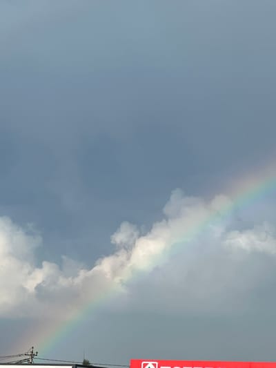 The image shows a wide shot of a cloudy sky with a faint rainbow visible on the right side. The bottom of the image features a section of a red sign that reads "TOTAL" in white lettering. A power pole with wires is visible on the left, and a few dark silhouettes of buildings and trees are present at the very bottom edge of the frame. The scene appears to be an outdoor setting, likely in a city or town, with overcast conditions. The location is identified as Mikhailovka, Russia.