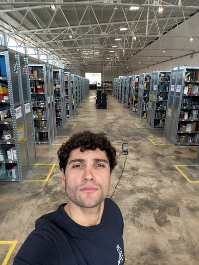 The image depicts the interior of a large industrial warehouse, identified as being in Polígon Industrial De Son Castello, Spain. A male subject is in the foreground, occupying the lower third of the frame, facing directly towards the camera. He possesses dark, curly hair, light skin, and facial stubble, and wears a dark blue t-shirt with a white owl logo and "NYC" text.
The background reveals a vast warehouse space characterized by numerous parallel rows of tall, galvanized metal shelving units extending into the distance on both the left and right sides of a central aisle. These shelves are stocked with a diverse array of items, including packaged goods, containers, and miscellaneous products. Specific shelf sections are labeled with white numbers such such as "34," "33," "19," and "20," and yellow adhesive labels, some displaying Spanish text like "ALQUILADO SEMANAL."
The floor is concrete, displaying scuff marks and tire residue, with yellow adhesive tape forming rectangular boundaries along the aisles. A small, black portable work light with a red electrical cord is visible on the floor in the mid-ground, slightly to the right of the central aisle. In the far distance, an individual wearing a high-visibility vest is partially visible among the shelves on the left side. A dark-colored, multi-tiered object on wheels, resembling a cart or stack of bins, is situated in the central aisle further back.
The warehouse ceiling is high and vaulted, featuring exposed metal trusses, white insulation panels, and multiple evenly spaced fluorescent or LED light fixtures. Rectangular skylights are also integrated into the ceiling structure, providing additional ambient light. The far end of the warehouse terminates in an open area that allows natural light to enter the facility.