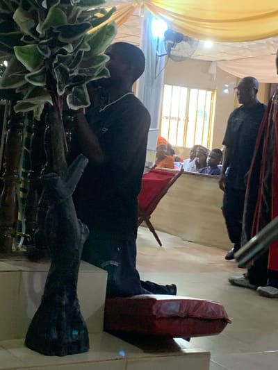 The scene appears to be indoors, possibly a church or a meeting hall, potentially in Akpaka Forest Reserve, Nigeria. The image is oriented sideways.
Several people are present. At least one individual in a black shirt is clearly visible, lying prone near some foliage and a dark, sculpted object (possibly a wooden carving or sculpture). Other individuals can be seen in the background, some wearing what appears to be traditional Nigerian clothing. There is also what appears to be a red cushion or bag near a white pillar.
The environment includes yellow and white drapery, a window with bars, and a fan. The lighting suggests an indoor setting. The overall atmosphere seems to indicate a gathering or ceremony.