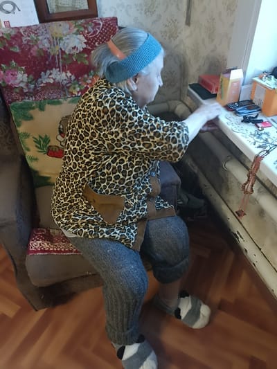 An elderly individual, identified as female, is seated in a fabric armchair in a residential room in Serpukhov, Russia. The individual has grey hair tied back with a light blue knitted headband and an orange hair clip, and is wearing a brown and black leopard print long-sleeved shirt with brown trim and pockets, grey ribbed trousers or leggings, and grey, black, and white striped slippers. The individual's right arm is extended, and their hand is resting on or reaching towards a white windowsill.
The armchair is covered with a patterned blanket or tapestry featuring red floral motifs and a beige section with a cartoon bear and green foliage. The windowsill is cluttered with various objects, including a red rectangular box, a cardboard box, two mobile phones (one flip phone, one smartphone), and several pieces of jewelry, among them a brown beaded necklace hanging over the edge. Below the windowsill, white heating pipes are visible. The wall behind the individual features light-colored patterned wallpaper. The floor is covered with light brown wooden laminate. A partial view of a mirror reflecting a white surface is visible in the upper left background.