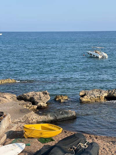 The image depicts a serene coastal scene in Antalya, Turkey, with the turquoise waters of the Mediterranean Sea meeting a rocky shoreline. In the foreground, a vibrant yellow dinghy rests on a pebble beach, its hull adorned with subtle yellow stripes. Beside it, a white surfboard lies partially visible, and a dark tarp covers another object. Further out in the water, a white motorboat with a canopy is anchored, its presence a solitary marker against the vast expanse of the sea. The rocky terrain bordering the beach is characterized by weathered stones and scattered debris, suggesting a natural and perhaps less manicured section of the coastline. The clear blue sky above, with a faint haze on the horizon, completes the tranquil Mediterranean vista.