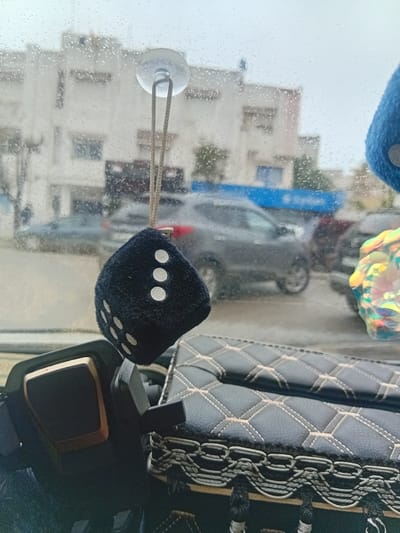 The image captures a view from inside a vehicle, looking out through a rain-splattered windshield into an urban or suburban setting in Martil, Morocco. The weather appears overcast and damp, with numerous water droplets visible across the glass, suggesting it is raining or has recently rained.
Inside the car, prominent objects include two fuzzy dice hanging from the windshield. A blue die is partially visible at the top, while a black die, showing faces with three and five white dots, hangs centrally from a white suction cup attached to the windshield via a cord. To the right, details of the car's interior are visible, including a grey or dark-colored quilted seat cover with light-colored stitching forming a diamond pattern, and a black and gold-trimmed car part, possibly part of the dashboard or a device. A colourful, iridescent, possibly holographic, ornament also hangs near the top of the windshield.
Through the wet glass, the blurred background reveals several light-colored buildings, likely residential, with visible windows. A dark-colored car is also discernable on the street or in a parking area, and a distinct vertical blue structure can be seen towards the left side of the exterior view. The scene is illuminated by daylight, but the overcast conditions create a soft, diffused light. There are no people visible, and no specific activity is taking place beyond observing the exterior scene from inside the car. No visible text is present in the image.
