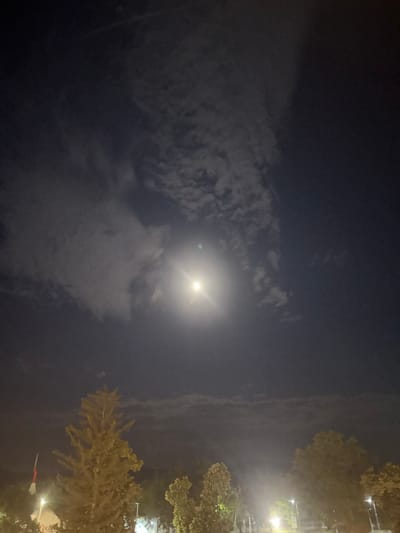This is a nighttime photograph taken in Kazanlak, Bulgaria, featuring a bright full moon partially obscured by clouds. The moon is the central subject, casting a luminous glow that illuminates the surrounding sky and creates a soft light. Wispy clouds drift across the frame, adding a dynamic texture to the dark sky.
To the right of the frame, the tops of trees are visible, their branches silhouetted against the night sky. In the lower right corner, what appears to be a flag or banner with red and white elements can be seen, possibly indicating a civic or national event. Streetlights or other artificial lights are also visible among the trees, suggesting an urban or park-like setting. The overall atmosphere is serene and a bit mysterious, characteristic of a moonlit night. The time of day is clearly night, and the presence of clouds suggests possibly a mild evening. No people are visible in the frame. No text is discernible in the image.