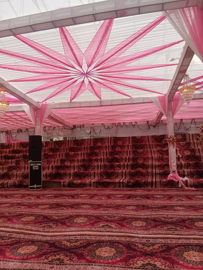 The image depicts an indoor event space, likely a venue for a celebration or ceremony. The ceiling is adorned with white and pink fabric draped in a starburst pattern, with small lights integrated into the pink fabric. Crystal chandeliers hang from the ceiling structure. Below the ceiling, tiered seating covered in a red and beige patterned carpet is visible. The carpet has intricate floral designs. On the left side of the frame, a black speaker is placed on the tiered seating, with text on its side reading "SAHI DJ & SOUND BADDI 9814140720". The overall impression is of a decorated venue prepared for an event. The location is identified as Manali, India, suggesting a cultural context for the decoration and setup. There are no people visible in the image.