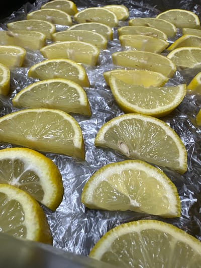The image displays a close-up, overhead view of numerous sliced lemons arranged in rows on a crinkled, transparent plastic sheet, likely plastic wrap. The lemons are cut into thin wedges, revealing their juicy interiors with visible segments and occasional seeds. The color palette is dominated by the bright yellow of the lemon rinds and the translucent yellow-green of their flesh. The lighting suggests an indoor setting, possibly a kitchen or food preparation area, with no distinct time of day or weather cues. The overall impression is one of food preparation or preservation, with the lemons laid out for drying, dehydrating, or as a garnish. There is no visible text in the image.