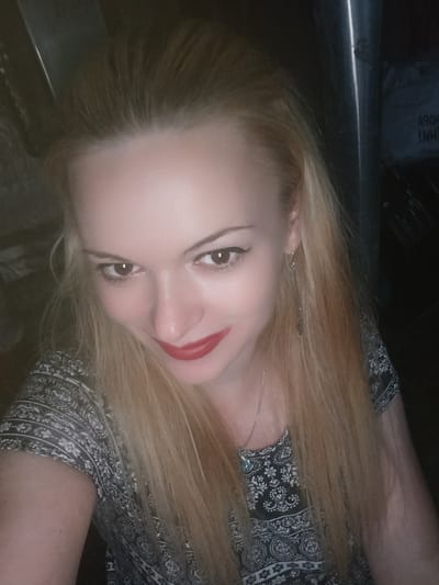 A woman with blonde hair and red lipstick is shown from the shoulders up, looking at the camera. She is wearing a black and white patterned dress and a silver necklace. The background is dark and out of focus, with a hint of a metallic pole on the right side of the image. There is text visible on a dark object in the background, which appears to be in an Eastern European language, possibly Ukrainian. There is no clear indication of her location within the city of Chernihiv, Ukraine.