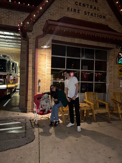 In front of the Austin Central Fire Station, a man in a white shirt and black pants stands next to a woman in a green jacket and jeans. A Dalmatian dog sits in a red wagon. String lights adorn the building's facade, and a fire engine is parked in the bay. Wooden chairs are stacked nearby.