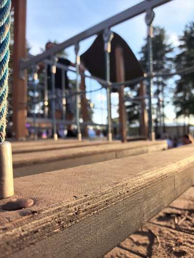 The image shows a close-up view of a wooden walkway or platform in a playground, with a rope in the foreground. In the background, a person is partially visible, blurred by the shallow depth of field. The playground structure appears to be made of wood and metal. The scene is set outdoors, with trees and a blue sky visible in the distance, suggesting it is daytime. The location is described as the city of Горный, Russia.