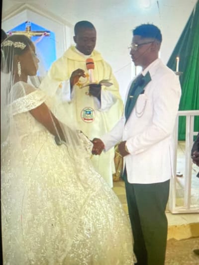 An indoor scene in the city of Mara, Nigeria, depicts a wedding ceremony involving three individuals positioned on a light-colored elevated platform. A dark-skinned woman on the left, identified as the bride, wears a white, off-the-shoulder lace wedding gown, a veil, and sparkling hair accessories. She faces a dark-skinned man on the right, identified as the groom, who is dressed in a white suit jacket, dark green trousers, a dark green ascot with a tie clip, and eyeglasses. The bride and groom are holding hands. Between and slightly behind them stands a dark-skinned man, identified as the officiant, clad in a light cream clerical vestment featuring an emblem. He holds a red-orange microphone in his right hand and a small open book in his left. The background features a blue crucifix on the left and a white candle on a stand partially visible against green drapery on the right.