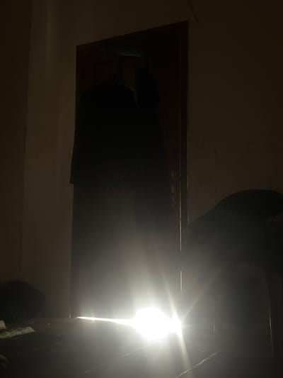 In Gaza City, Palestinian Territories, a dimly lit room features a door ajar, with a bright light emanating from the gap at the bottom. A dark silhouette of clothing hangs on the door. The floor is visible with scattered objects. A dark, indistinct shape, possibly furniture covered with cloth, is visible to the right. The walls of the room are illuminated by the light from the doorway, casting long shadows.