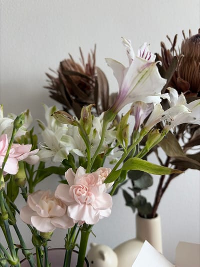 The image displays a floral arrangement set indoors in Yanino-1, Russia. The foreground features a bouquet predominantly composed of light pink carnations and white alstroemeria with green stems and buds. Some alstroemeria flowers exhibit faint purple and green striations. In the background right, a separate arrangement consists of multiple dark brown, dried, spiky botanical elements contained within a slender, light beige ceramic vase, partially obscured. A clear, possibly purple-tinted, glass vase is partially visible at the bottom left, holding some fresh flowers. Ancillary objects on the surface include a light-colored, animal-shaped ceramic figurine, another rounded light-colored ceramic item, and a white piece of paper, all positioned against a plain, light-colored wall. No individuals are present.