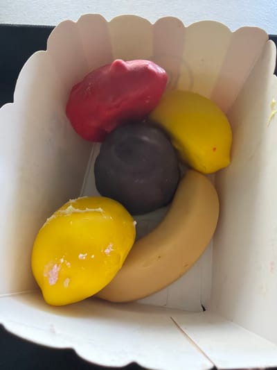 A top-down, medium shot captures a close-up view of five colorful, food-shaped candies nestled within a white, fluted paper container, likely a dessert cup. The candies are arranged in a loose cluster. At the top left, a bright red, slightly irregular oblong candy sits. Adjacent to it, towards the right, is a bright yellow, elongated candy with a small red dot near its tip. Below the red candy, a dark, almost black, round candy is positioned centrally. To the left and slightly below the dark candy, another bright yellow candy, also elongated, is visible, with some white markings on its surface. Finally, a pale yellow, banana-shaped candy curves along the bottom right side of the container. The paper container has a scalloped edge and appears to be made of a stiff, textured material. The background is out of focus, showing a hint of a dark surface and a light, textured wall or surface. The lighting is bright, casting subtle shadows. No geographical location can be determined from the image.