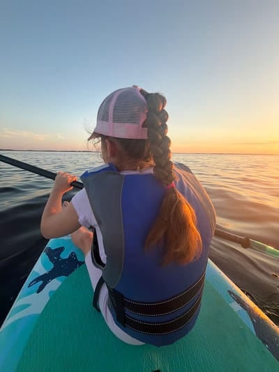 A young girl with a braided auburn ponytail, wearing a pink and white trucker hat and a blue and gray life vest, sits on a turquoise stand-up paddleboard. She is holding a paddle, its shaft black and its blade partially visible and green. The paddleboard is on dark water with visible ripples. The background shows a calm body of water extending to the horizon, meeting a clear sky. The setting sun casts a warm, orange glow across the sky and reflects on the water, creating a serene sunset scene. The image appears to be taken during the golden hour.
