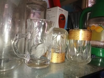 A close-up, indoor still life shot depicts an arrangement of glass and household objects on a reflective dark surface. From left to right: a tall, cylindrical clear glass container stands prominently. Adjacent to it, centrally positioned, is an empty clear glass mug featuring an ornate handle and frosted leaf patterns etched into its side. To the immediate right of the mug, an empty clear glass tumbler displays a wide gold-colored decorative band around its upper rim and a faceted lower body. Behind this tumbler, a partially obscured clear glass object, possibly inverted, shows a gold-patterned band near its visible base. On the far right, a green plastic thermos or pitcher with a ribbed transparent section is decorated with yellow fruit decals, likely lemons. In the upper left background, a white cardboard box labeled "ADVANCE Bluetooth Speaker" with an image of an orange speaker is visible. Dark, indistinct objects are discernible in the background behind the central glass items. The scene is static, devoid of people or ongoing actions. This scene is located in Emthanjeni Local Municipality, South Africa.