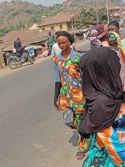 The image portrays a daytime street scene in Ogaminan, Nigeria. A paved road occupies the foreground, extending into the midground where it is bordered by buildings and individuals. In the center-left foreground, a young adult female stands, looking directly at the camera. She wears a black headscarf, a light blue collared shirt with green, orange, and grey abstract patterns, matching patterned trousers, and long black undersleeves. Dark sandals are visible on her feet. To her right, the back of another individual, likely female, is partially visible, wearing a black head covering and a vibrant blue, orange, and green tie-dye patterned dress or wrapper.
Behind these two figures, a small group of people is present. One person wears a black cap, and another has a multi-colored patterned headwrap (red, black, white). On the left side of the midground, a silver motorcycle, equipped with a blue and white patterned box on its rear seat, is parked. A man in dark blue attire stands near the motorcycle, facing away from the viewer. Further back, a man in a green sleeveless top and trousers is visible near buildings. Several large white sacks are resting on the ground in this vicinity. The background features residential-style buildings with light-colored walls and brown corrugated roofs, situated at the base of arid, sparsely vegetated hills under a clear sky.