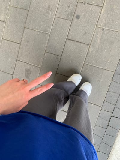 A person is standing on a grey concrete sidewalk in Kraków, Poland, at the location of latitude: 50.068, longitude: 19.949. The person is wearing a blue shirt, grey pants, and white shoes. The person is making a peace sign with their right hand, which is visible in the image. The person's left hand is not visible. The concrete sidewalk is made of square tiles. The person is standing in a slightly angled position. The person is facing away from the camera. The person is in a casual, relaxed pose. The background is a blur of grey concrete tiles. The overall scene is simple and mundane.