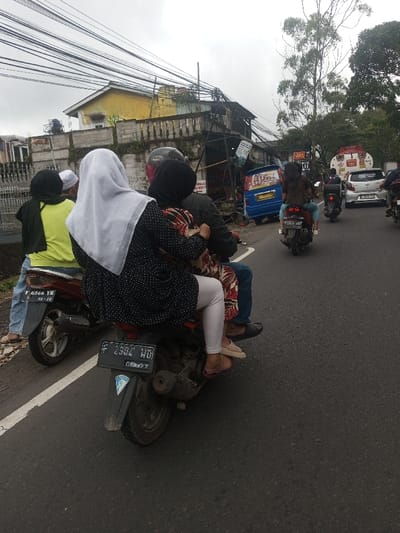 A motorcycle with three occupants travels on an asphalt road. The passenger in the rear wears a white hijab, while the middle passenger has a dark headscarf. Several motorcycles are ahead in the same direction. Buildings line the side of the road, including one with a yellow facade. Overhead are numerous electrical wires. A large tanker truck is visible in the distance. The sky is overcast, suggesting a cloudy day.