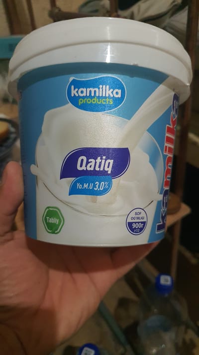 A person is holding a white plastic tub of "Qatiq" (yogurt) with a blue and white label in Karankul, Uzbekistan. The label features the brand "Kamilka Products" in blue and white lettering. The tub indicates "Yo.M.U 3,0%" (fat content) and "Sof Og'irligi 900r ±10" (net weight). A green oval sticker with "Tabiiy" (natural) is also visible on the tub. The background is blurred, showing indoor elements possibly in a home or store setting.