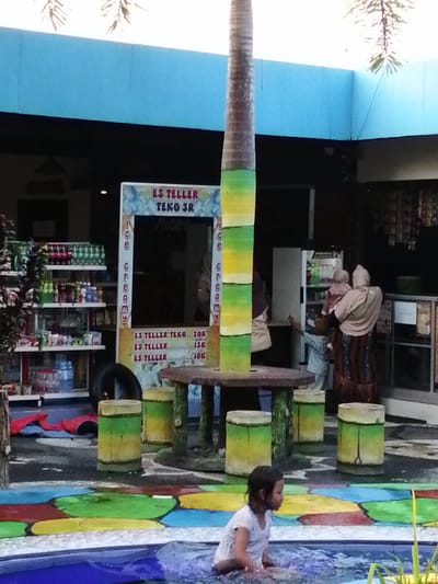In Kebonsari, Indonesia, a young girl in a white t-shirt sits in a blue pool. Behind her, a cylindrical object, possibly a decorative tree, has sections painted in green and yellow stripes. To the left, shelves are stocked with beverages. In the center, a food stall displays prices for "ES TELLER" (ice teller), with options ranging from 10K to 20K. In the background, several individuals are gathered near a refrigerator, suggesting a retail or food service environment. The area appears to be an outdoor recreational space with colorful, patterned flooring around the pool.