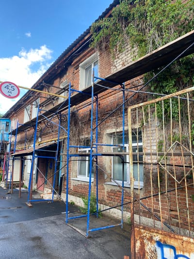 The image depicts an urban scene with scaffolding erected against a weathered brick building, suggesting ongoing construction or repair work. A street sign indicating a height restriction of 4.5 meters is visible, suspended by yellow poles. The building shows signs of age, with vines growing on its facade. A rusty metal gate stands in the foreground. The ground appears wet, indicating recent rainfall. The overall impression is one of a slightly neglected but functional urban environment. This scene is located in the city of Samara, Russia.