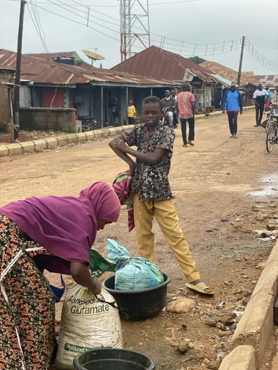 The image shows a street scene in Kafanchan B, Nigeria. A young person wearing a dark patterned shirt and yellow pants is standing and smiling at the camera. Another person, wearing a purple head covering and a dark floral-patterned dress, is bending over a large white bag with "Monosodi Glutamate" printed on it. Next to the bag are two black buckets. In the background, other people are walking along the dirt street. There are buildings made of corrugated metal and a communications tower visible in the background. The ground is dirt and littered with debris.