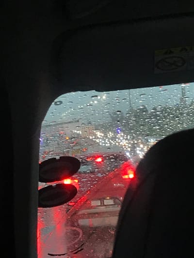 The image provides an interior view from a vehicle, looking forward through a heavily rain-streaked windshield. The scene depicts a densely congested urban road during heavy rainfall, likely at dusk or night, in Sandton, South Africa.
In the immediate foreground, dark interior elements of the vehicle are visible, including the upper-left A-pillar, a portion of the dark headliner, and the upper-right part of a dark seat headrest. Two circular, dark-colored suction cups are affixed to the lower-left interior of the windshield. A yellow warning label, possibly related to an airbag, is partially visible on the passenger side sun visor in the upper-right quadrant.
The midground and background are dominated by numerous vehicle lights, heavily diffused by the raindrops on the windshield. Bright red taillights from multiple vehicles are prominent in the immediate road ahead, indicating stopped or slow-moving traffic. Beyond these, numerous white lights, likely headlights from distant traffic or streetlights, are visible. A hazy urban skyline with discernible building structures and scattered illumination is present in the far background. The overall environment is characterized by adverse weather, poor visibility, and significant traffic congestion within a developed city setting.