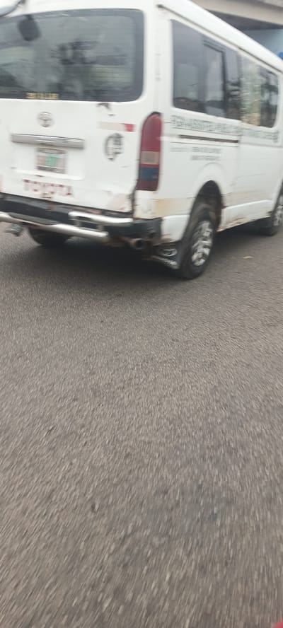 The image shows the rear of a white Toyota Hiace minivan driving on a paved road. The van is dirty, with mud splattered on its lower panels and rear bumper. A chrome bumper guard is installed, and dual exhaust pipes are visible. The license plate is partially obscured but appears to have green characters. The van has a rear window with a wiper. Text and logos are visible on the van's body, including a Toyota emblem and what appears to be an inscription on the side. The scene is set in the city of Kabong, Nigeria, characterized by the worn asphalt road and the type of vehicle commonly used for public transportation or commercial purposes in the region. The image is slightly blurred due to motion.