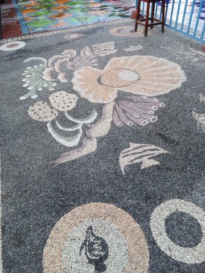 The image displays a mosaic pathway or floor made of small pebbles embedded in cement. The mosaic depicts various marine-themed elements, including a large seashell, stylized coral or seaweed formations, a dolphin, and other abstract shapes resembling underwater flora. The pathway is dark gray with lighter gray pebbles forming the intricate designs in colors of white, beige, pink, and green. In the background, a colorful tiled area, possibly a pool deck, is visible, with reflections suggesting it is wet. A wooden stool and a blue metal railing are also present, indicating an outdoor setting, likely near a body of water. The overall scene suggests a decorative outdoor space in Kebonsari, Indonesia.