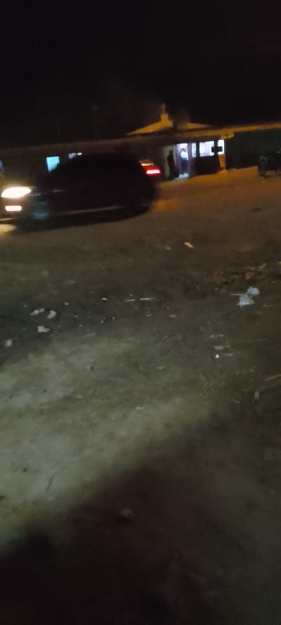 A dark, blurry nighttime scene features a car with illuminated headlights and taillights parked on a rough surface with scattered debris. In the background, a building with lit windows has a person standing near the entrance. A motorcycle is also visible. The overall image quality is poor, with low light and significant blurriness.