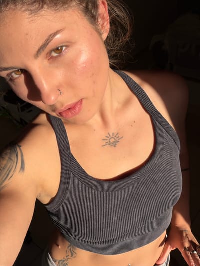 A young woman is shown in a close-up, angled, eye-level shot. She has fair skin and light brown hair. Her eyes are hazel-colored, and she has a nose piercing. She is wearing a gray ribbed tank top. A tattoo of a sun is visible on her chest. Another tattoo is partially visible on her arm. The lighting is bright and casts shadows across her face and body. No location information in the city of Kiryat Bialik, Israel is visible in the image.