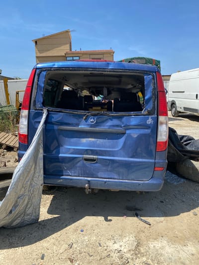 The image shows the back of a damaged blue Mercedes-Benz Vito van in what appears to be a junkyard or salvage yard. The van has significant damage to the rear, with the rear window entirely missing, along with dents and damage to the body. The van is located in Baraki, Algeria.
The environment surrounding the van consists of a concrete and dirt surface, with other vehicles and debris visible in the background. There are no people visible in the image.