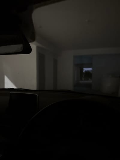 The image presents a static, low-light scene captured from inside a vehicle, located in Latsia, Cyprus. The lower half of the frame shows the vehicle's dark dashboard, with a black rectangular display unit positioned centrally on the left side, and a portion of a dark steering wheel visible on the lower right. The upper left reveals a segment of the rearview mirror assembly. Beyond the windshield, a dimly lit interior structure with white or light grey walls is visible. On the left, a bright triangular section suggests an angled wall or light reflection. In the mid-ground, a series of dark rectangular openings or doorways lead into a further space, characterized by light blue-grey walls. Through a distant rectangular opening within this space, an exterior environment, appearing to be night or heavily shadowed, shows indistinct dark shapes possibly representing foliage. To the far right, a large, white cylindrical object, consistent with a water tank or similar container, is partially discernible against a wall. No individuals or specific actions are depicted within the frame.