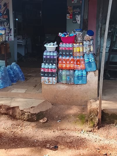 The image shows a street-level view of a small shop in Kabong, Nigeria. In the foreground, a sandy and uneven dirt ground is visible, with some scattered debris and small green plants. A low, rough, concrete structure forms the base of the shop's display area.
On this concrete base, an assortment of bottled beverages and food items are stacked. To the right, there are several large blue plastic bottles of water, some stacked in pairs and others individually. To the left of these, there is a stack of smaller plastic bottles containing dark-colored soda, some with red labels that appear to be Coca-Cola. Above these, there is a layer of orange-colored soda bottles, possibly Fanta, and a stack of what seem to be multipacks of canned beverages.
At the top of the beverage stack, a pink plastic bowl holds an assortment of snacks, likely chips or crackers. Behind this, a large clear plastic container with a blue lid is filled with what appears to be nuts or other dry goods.
Further back, the entrance to the shop is visible. It is dimly lit and appears to be a simple open doorway. A poster with text and images, including what looks like "EWA STUDIO," is mounted on the wall to the left of the entrance. Inside the shop, some furniture and equipment are vaguely discernible in the shadows. The overall impression is that of a small, informal retail establishment catering to local needs, common in areas like Kabong.