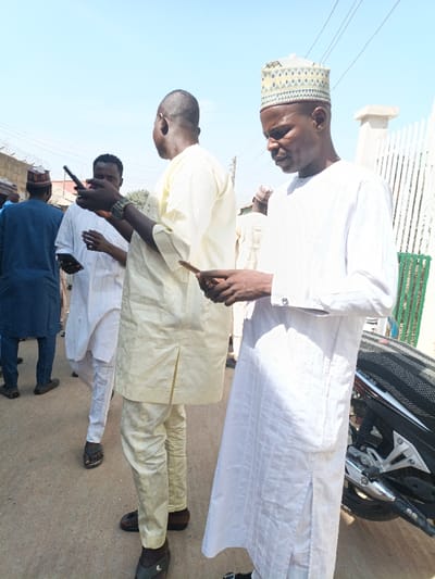 The image depicts an outdoor scene in Gawuna, Nigeria, under bright daylight with a clear, light blue sky. At least five adult males are present, primarily focused on handheld mobile phones.
In the foreground, a man on the far right wears a white, long-sleeved traditional tunic with subtle textured patterns and a patterned light-colored cap. He looks downwards, holding a dark-colored mobile phone horizontally with both hands. To his left, another man with a dark complexion wears a light yellow long-sleeved traditional tunic and matching trousers. He faces slightly away from the camera, holding a dark mobile phone vertically with both hands, also looking at it. A dark watch is visible on his left wrist. Further left in the mid-ground, a man with a dark complexion wears a white long-sleeved traditional tunic with vertical stripes and matching trousers, holding a dark mobile phone in his left hand while looking at it.
In the background, two additional men are visible: one wearing a dark blue traditional tunic and cap, with his back to the camera on the far left, and another partially obscured figure.
The ground surface is a light-colored, possibly dirt or concrete pathway. On the far right, a portion of a black and silver motorcycle is visible, including its textured seat and engine components. The background features a white wall with a vertical white metal fence or gate on the right, and a lower, light-colored wall topped with barbed wire on the left, indicative of property boundaries. Utility lines are visible overhead against the clear sky.