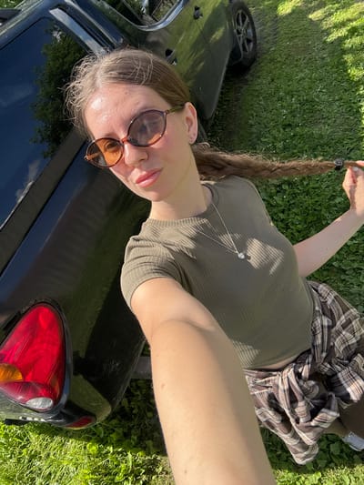 A woman is taking a selfie outdoors, leaning against the side of a dark-colored vehicle. She is wearing a green ribbed t-shirt, a plaid shirt tied around her waist, and sunglasses with oval, amber-tinted lenses. Her hair is long and braided. The vehicle appears to be a pickup truck, with its rear taillight visible. The ground is covered in green grass, suggesting a rural or park-like setting. The lighting indicates it is daytime. The woman is holding a strand of her braid with her right hand. There are no other people or animals clearly visible in the frame. The background shows more green grass and some trees.