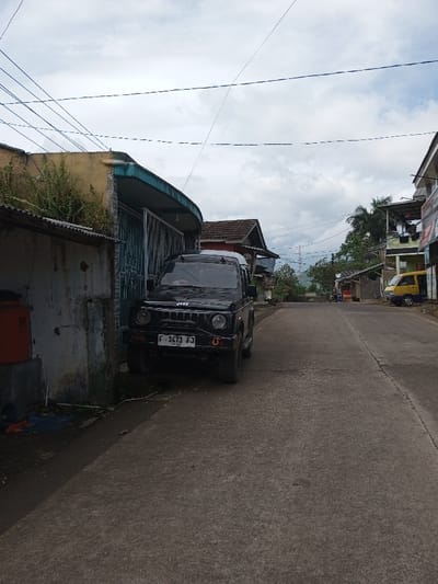 The image depicts a daytime street scene identified as being in the city of Ariyalai, Sri Lanka, under an overcast sky. A black four-wheel-drive vehicle, resembling a Suzuki Jimny, is prominently parked facing the viewer on the left side of a concrete road. The vehicle displays an Indonesian license plate, F 1473 FJ, affixed to its front bumper. To the immediate left of the vehicle is a weathered building featuring a curved, teal-colored awning above a metallic gate. An orange water tank rests on a concrete base against the building's wall in the foreground. Overgrown green vegetation is visible around the building and fence line.
The concrete road, which appears worn with visible seams, extends uphill and curves slightly to the right in the distance. On the right side of the road, various single and multi-story buildings line the street, some with visible commercial signage. A yellow van is parked further down the road on the right. Lush green trees and foliage are present behind the buildings, leading to distant hills or mountains visible under the cloudy horizon. Multiple black power lines crisscross the sky overhead. No individuals are present, and no active events are occurring within the visible scene.