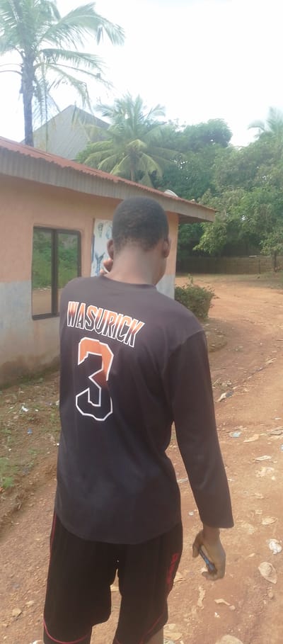 In this medium shot, a young Black male is captured from the back, walking away from the camera on a dirt path. He is wearing a long-sleeved black t-shirt with "WASURICK" and the number "3" prominently displayed in orange and white lettering. He is also wearing black shorts with red trim. The man is holding a blue mobile phone in his right hand, suggesting he might be on a call or using it.
The setting appears to be a rural area in Achara Ndiagu Omegio, Nigeria, with a modest, light-orange building to his left, featuring a single window and a corrugated metal roof. Palm trees and other lush green foliage surround the building and extend into the background, indicating a tropical or subtropical environment. The sky is overcast and bright, suggesting it might be daytime, possibly after a rainfall or with impending rain, as the ground appears damp and dusty in places. The overall atmosphere is casual and unpretentious, a snapshot of everyday life in a residential, possibly semi-urban or rural, Nigerian landscape.