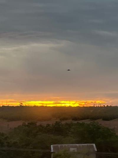 An image shows a sunset over Karagash, Moldova. The sky is a mix of gray and orange clouds, with the sun casting a bright yellow glow on the horizon. A small, dark object, possibly an aircraft, is visible in the upper part of the sky. Below the sky, a dark silhouette of trees and hills is visible, with a structure that appears to be a building roof in the foreground. The overall impression is a peaceful evening scene in a rural setting.