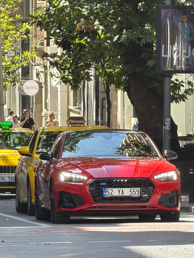 In Şişli, Turkey, a bright red Audi sedan is parked on a city street, its front prominently displayed with a Turkish license plate reading "52 YA 559." The car is positioned behind a yellow taxi, a portion of which is visible on the left. The street appears to be lined with buildings, with trees and foliage casting shadows. In the background, a woman with blonde hair is visible, along with other individuals, some of whom appear to be pedestrians. A circular sign with the word "HOTIC" is also visible on a building to the left. The scene suggests a sunny day in an urban environment, likely a commercial or residential district.