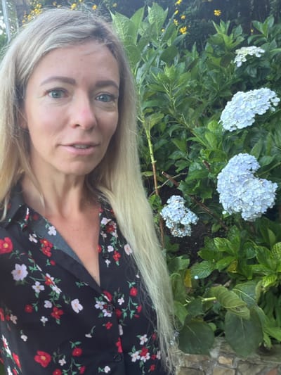 A woman with blonde hair is taking a selfie in front of a bush with light blue hydrangeas in Xuân Thành, Vietnam. She is wearing a black collared shirt with a red and white floral pattern. Her hair is down and covers her left shoulder. The background is filled with lush green foliage and the pale blue flowers.
