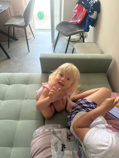 An indoor scene shows two children on a light green tufted sofa. The child on the left is a blonde toddler wearing a pink top, facing the camera with wide blue eyes and a slight smile. The child on the right is partially visible, wearing plaid shorts and a white t-shirt with a design. This child's arm is outstretched, holding a stylus over a tablet device. In the background, through a glass door, a balcony is visible with a green and blue inflatable ball. Two chairs are also visible; one is dark gray with black legs, and the other is lighter gray with a white base. Clothes are draped over the dark gray chair. The overall environment suggests a casual domestic setting. The specific location of Novorossiysk, Russia, is not discernible from the visual information provided in the image.
