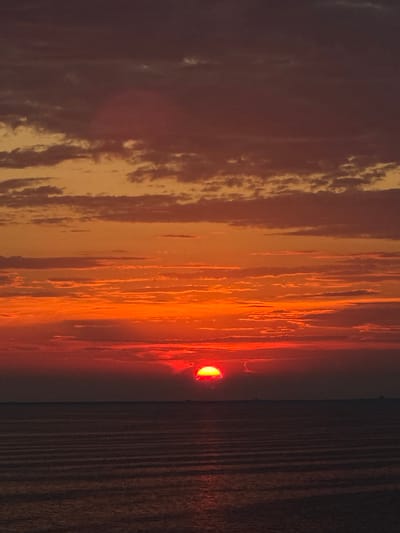 The image depicts a sunset over a body of water, possibly the sea. The sky is filled with vibrant hues of orange, red, and dark purple, with scattered clouds. The sun, a bright red orb, is partially obscured by clouds near the horizon. The water below is dark and appears to have gentle ripples. There are no discernible people, objects, or specific landmarks that would indicate a location within a city, whether in Italy or elsewhere. The scene is a natural landscape view.