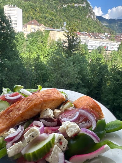 A close-up shot of a white plate containing a salmon salad with feta cheese, cucumber, red onion, tomato, and green bell pepper, set against a blurred background of trees, buildings, and a mountain. The buildings appear to be in the city of Saint-Claude, France, a town nestled in the Jura Mountains. One tall, modern building is visible on the left, while older, traditional buildings with red-tiled roofs are seen further in the background. The mountain slope behind the town is covered in dense green trees, with a rocky, white cliff face visible near the top under a blue sky with scattered white clouds. The lighting suggests it is daytime, likely late morning or early afternoon. There are no people visible in the image.